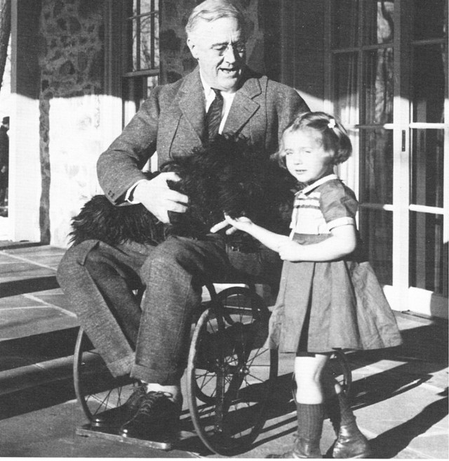 A wheel-chair bound FDR holds a dog in his lap and interacts with his granddaughter. 