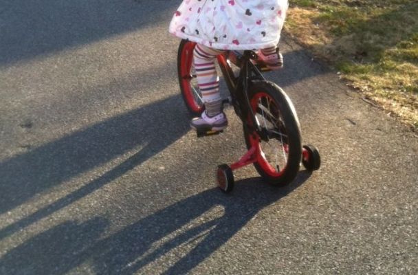 A four-year-old girl in a polka dot jacket peddles down the sidewalk on a tiny red bike with training wheels.