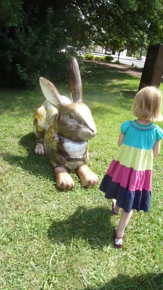 A five year old girl stands in a park facing a large brown painted rabbit. 