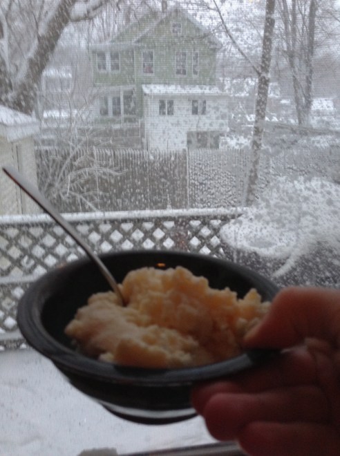 A hand holds a bowl of what looks like vanilla ice cream in front of a snowy window. 