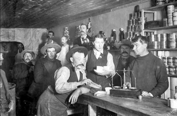 In this black and white photo, miners wait as the store manager weighs out their gold dust.