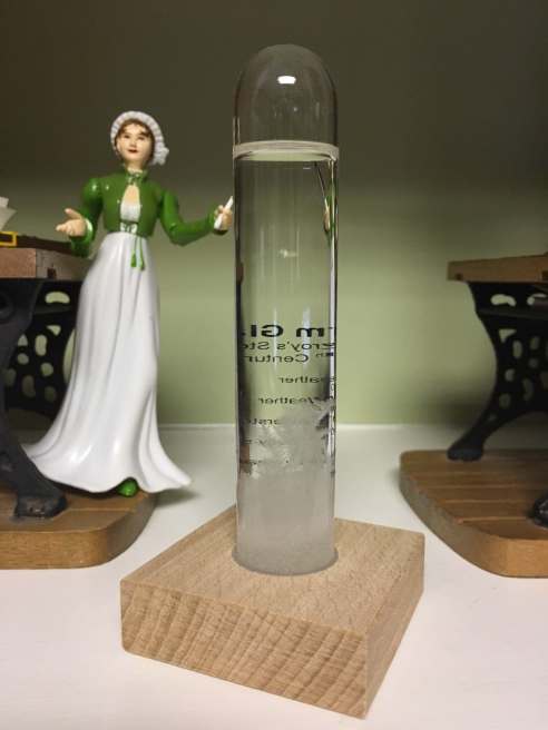 Photo of a Jane Austen action figure posed on a bookshelf next to the no longer working storm glass.