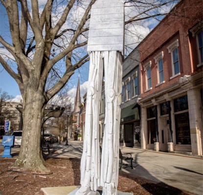 Photo shows a house perched on very thin, very tall, vaguely robotic sculpted legs in front of a brick building.