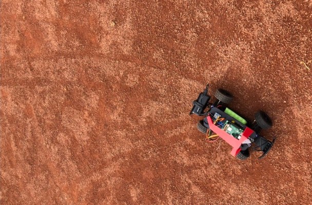 View of a remote control car from above, showing the tracks the car's tires have left on the bright orange dirt of a baseball field.