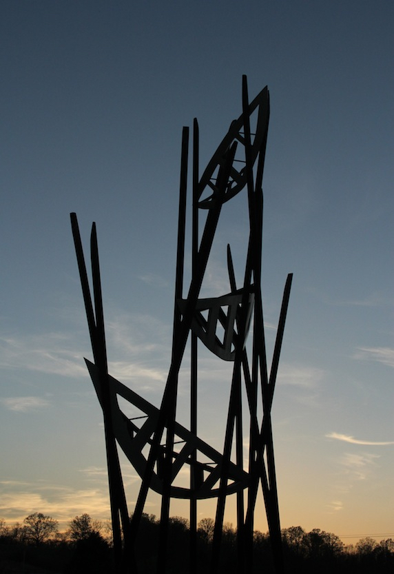 The sculpture resembles a series of three open work canoes suspended at various heights on a network of angled poles. The blues, greys, and orangey-pinks of the evening sky show through the sculpture. 