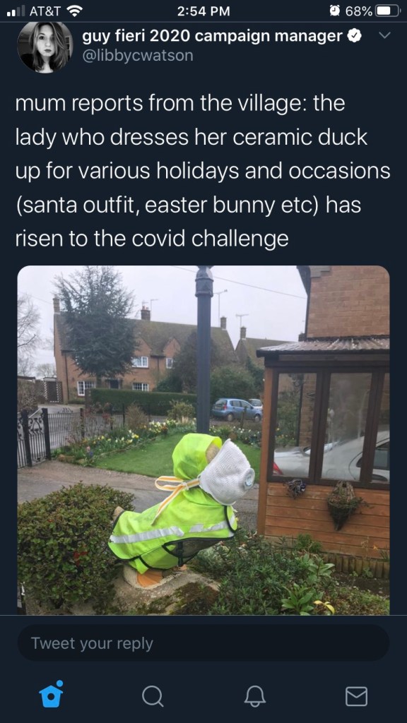 Tweet from @libbycwatson reads: "Mum reports from the village: the lady who dresses her ceramic duck up for various holidays and occasions (santa outfit, easter bunny etc) has risen to the covid challenge." Image is of a duck status in a garden covered in a crossing guard's uniform and wearing a face mask (hopefully not a medical grade one). 