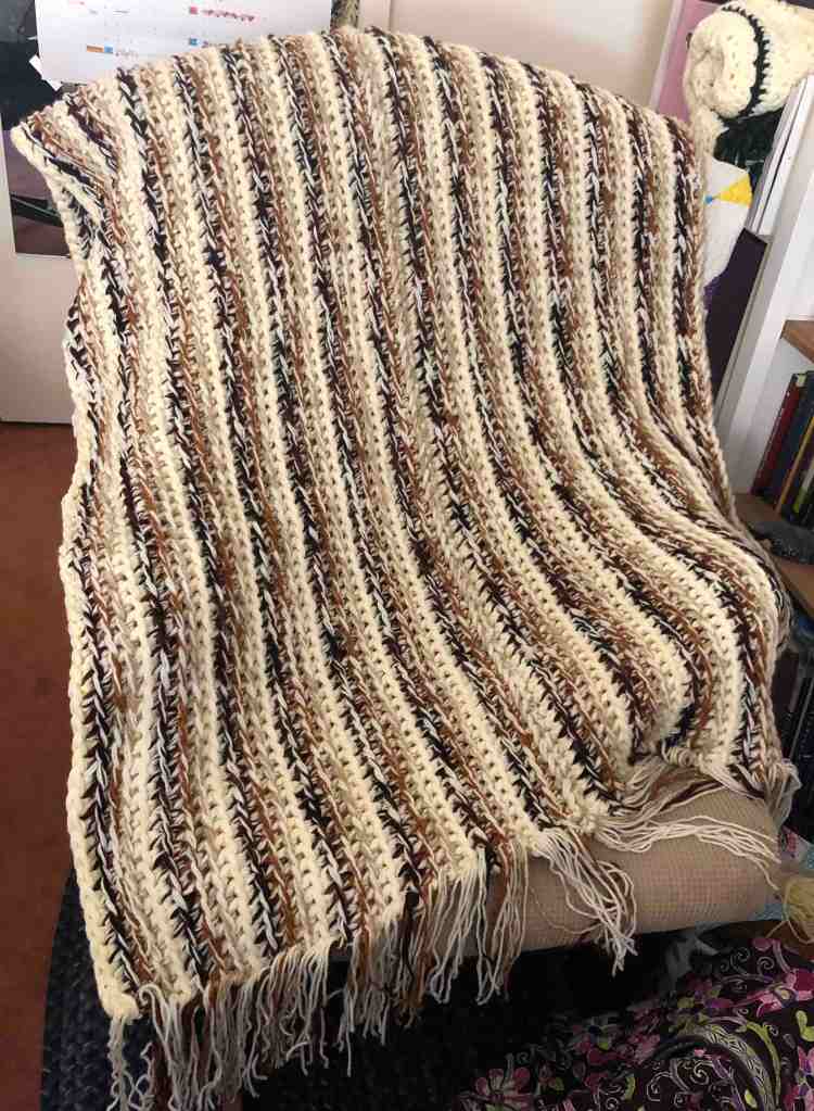 Photo shows a crocheted striped blanket draped across a rocking chair. The blanket is striped and crocheted in cream, light brown, rusty brown, and dark brown yarns.