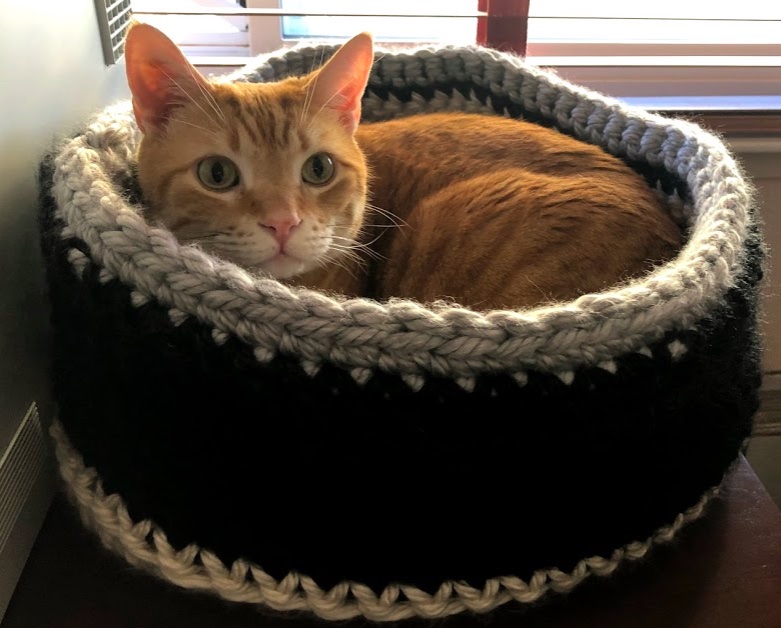 Orange cat nestled in a black, grey, and cream cat bed crocheted out of insanely bulky yarn. 