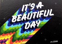 Mural displays the words It's a Beautiful Day painted in white in a rounded all-caps font against a black background. The artist has painted a multi-layered shadow for each of the letters in the words out of the colors of the rainbow. The effect is to make the words look like they've been launched into space, trailing rainbow flames. (Sorry this is a terrible description.)