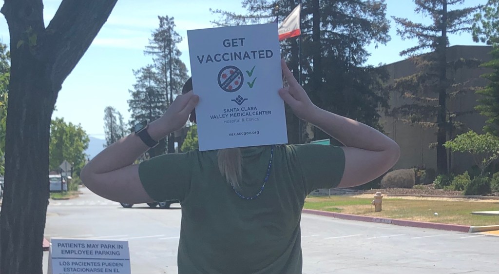 My daughter holds a sign from the vaccination center in front of her head. The sign reads "Get Vaccinated! Santa Clara Valley Medical Center". She is standing in a parking lot with pine trees and the California state flag visible behind her.