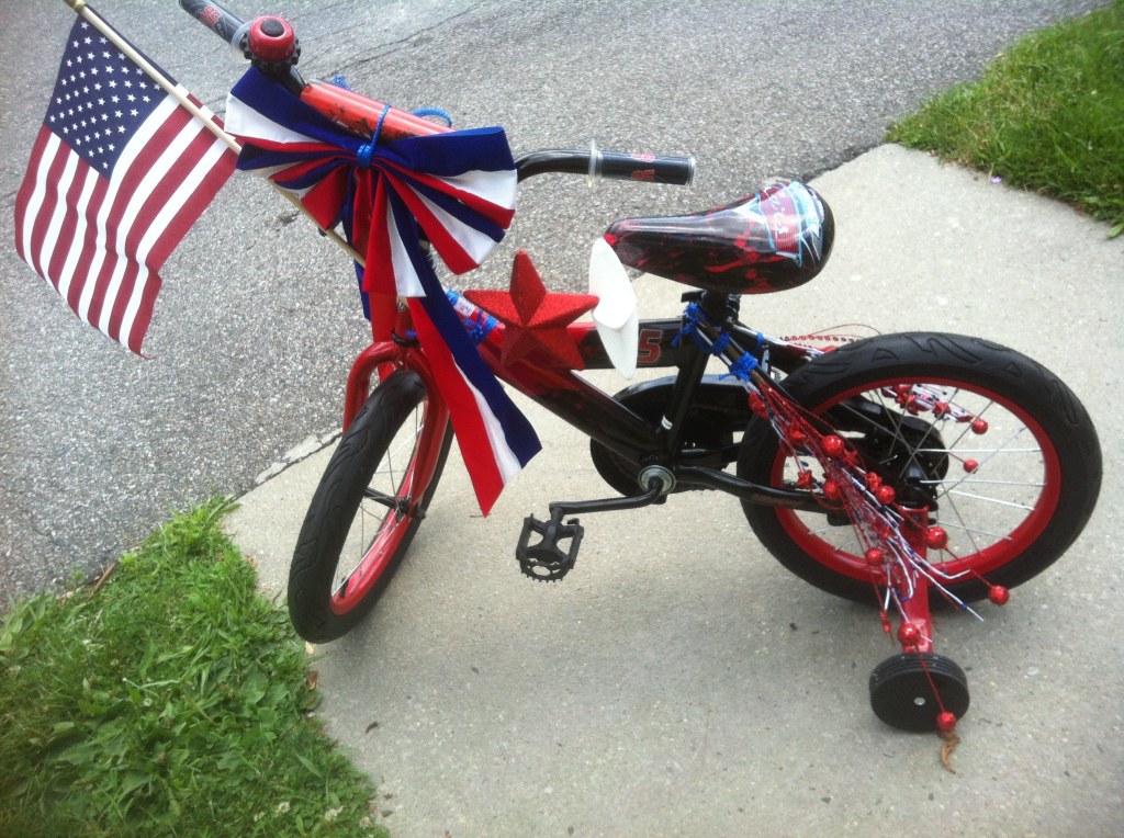 Red bike with training wheels, dressed up for the holiday with an improbable number of flags, stars, oversized red white and blue bows, and plastic fake fireworks. 