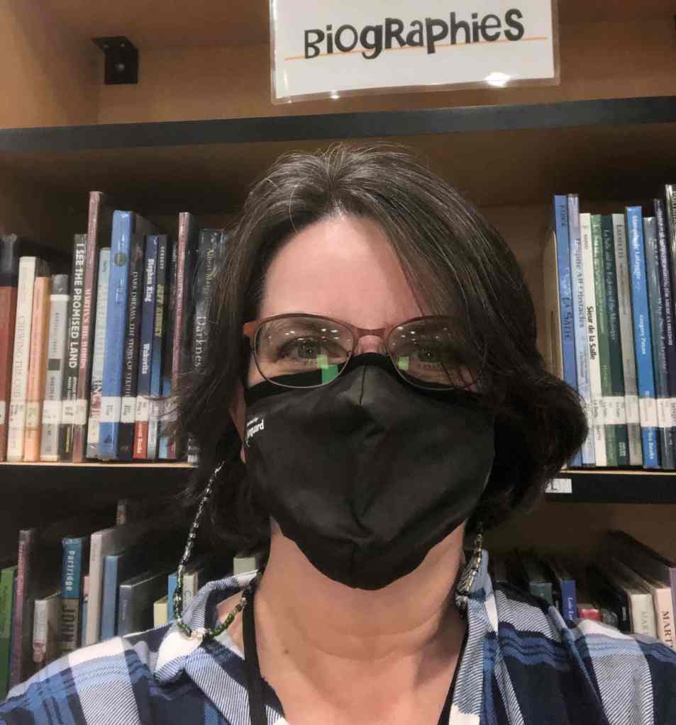 Me, with short hair and mask, standing in front of one of the biography stacks at the middle school library. 
