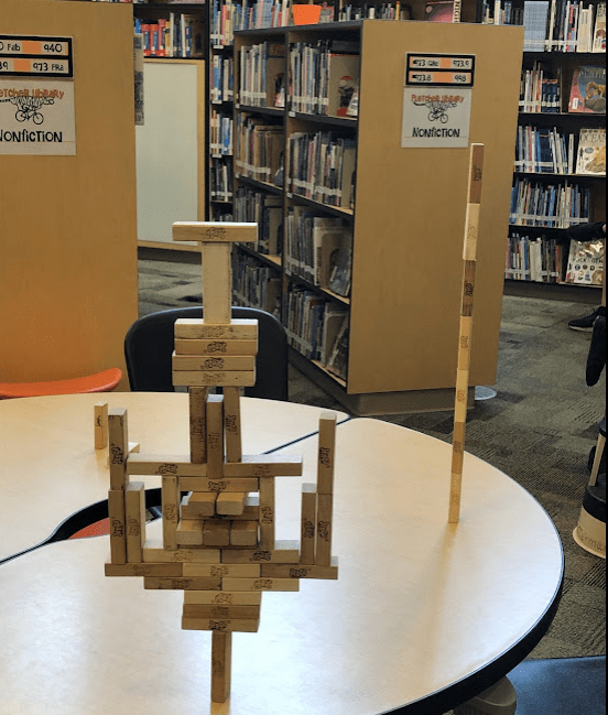 In the foreground, you see an improbably balanced sculpture made of Jenga blocks on a round table. Behind it are some of our nonfiction stacks, which consist of rows of densely packed, spine-out books with an occasional forward-facing book.