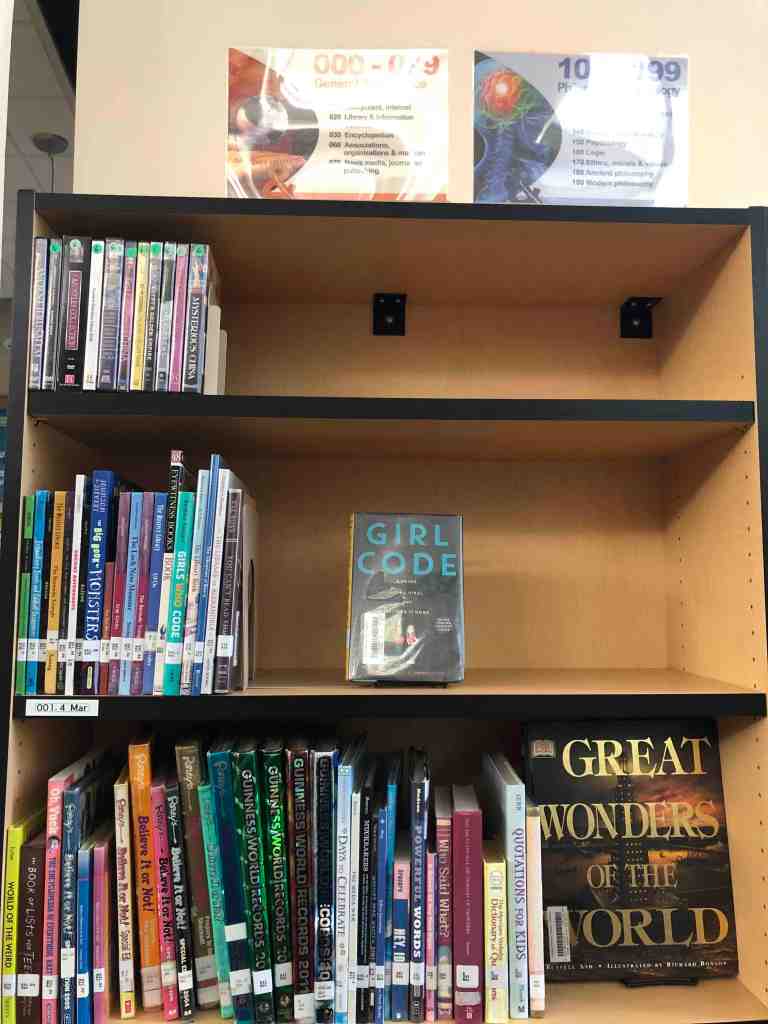 Photo of the nonfiction stacks with the original nonfiction signs. There is a glare obscuring much of the sign, but the sign for 000-099 reads General Reference and lists the major topics by decade (eg. 030 Encyclopedias). 