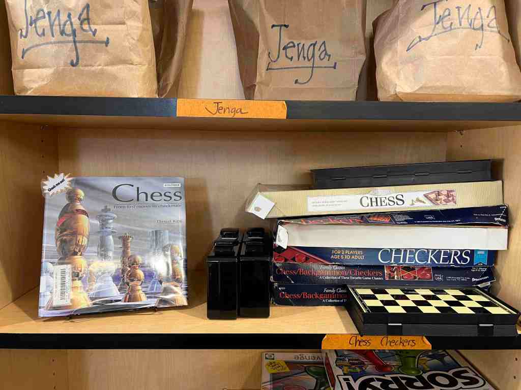 Photo of our games shelf. Jenga on the shelf above, chess & checkers on the middle shelf, with a book on Chess on display next to them. 