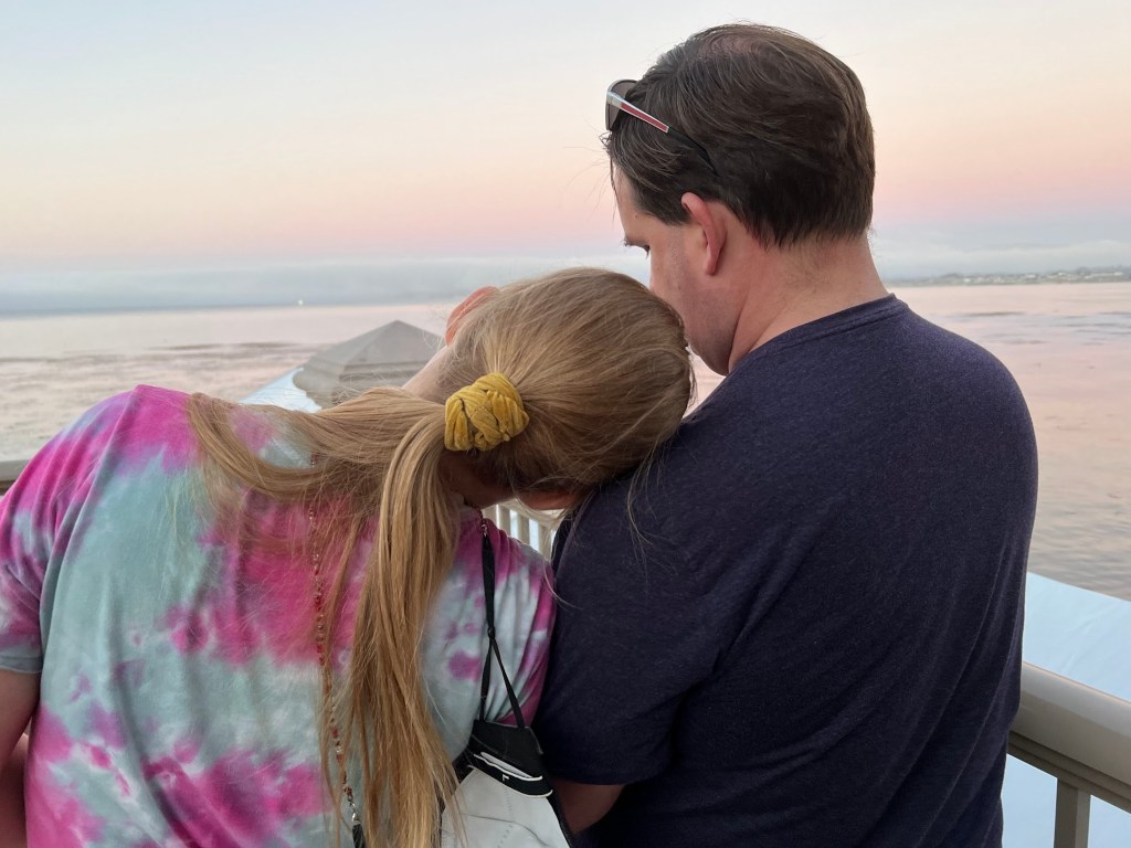 Image shows my husband and daughter watching the sunset together. The picture is taken from behind. My daughter is resting her head on her dad's shoulder. 