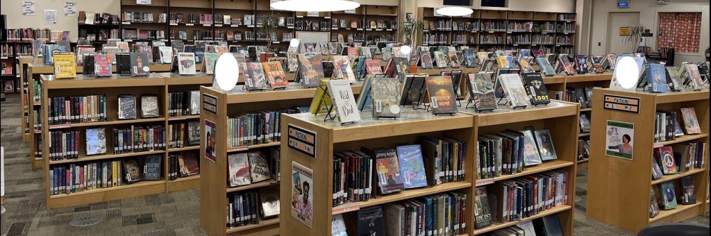 image shows a wide angle view of the library where I worked in May 2023. The stacks in front are half-sized, with books arranged in a display on top of each stack, and more positioned front facing on the lower shelves to encourage browsing.